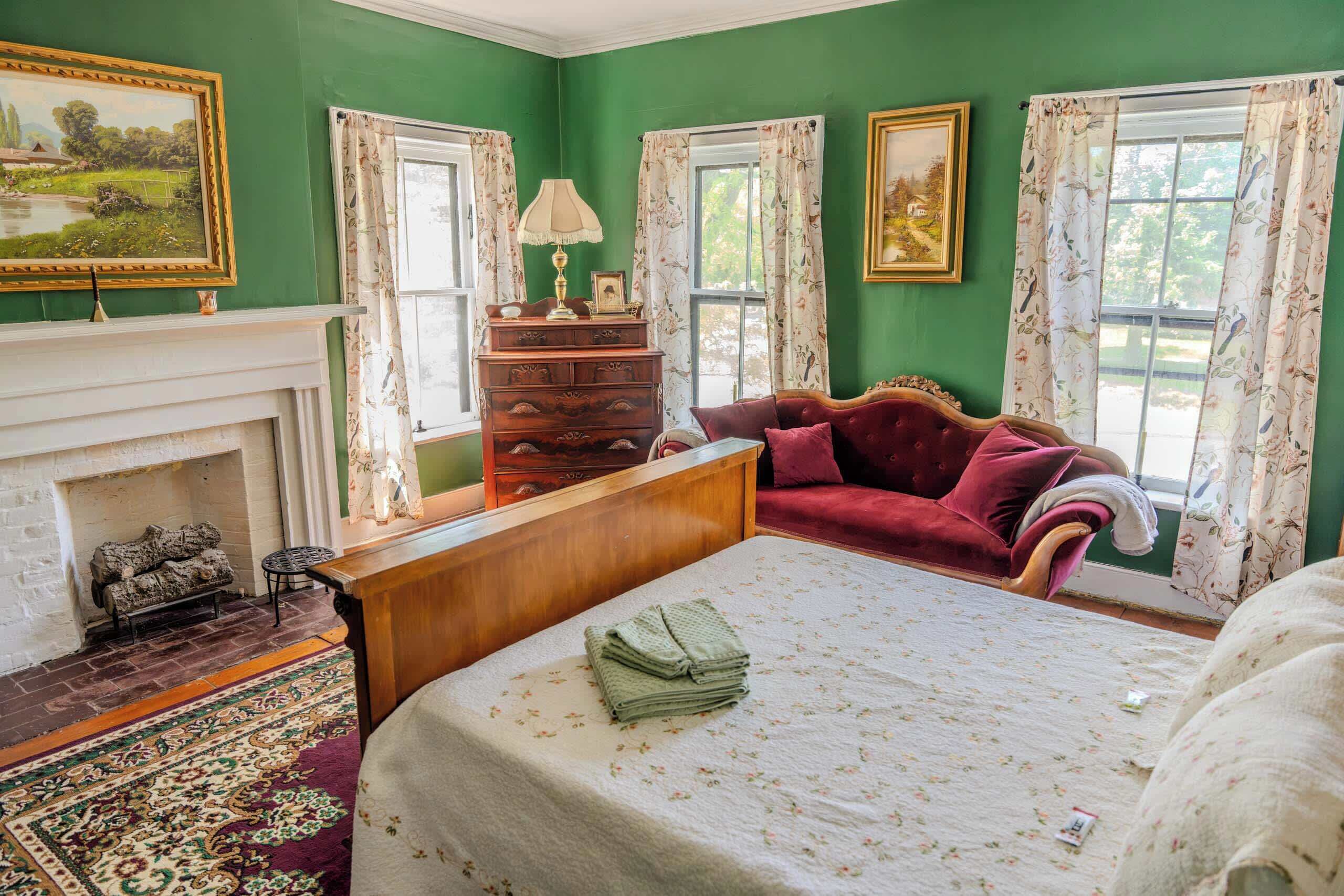 A room with green walls, a fireplace, a bed with a floral bedspread, a red sofa, and two windows with floral curtains.
