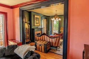 A room with a fireplace, a vintage armchair, a chest of drawers, and a large window with a view of greenery. - The Gould Residence (Seneca Falls)