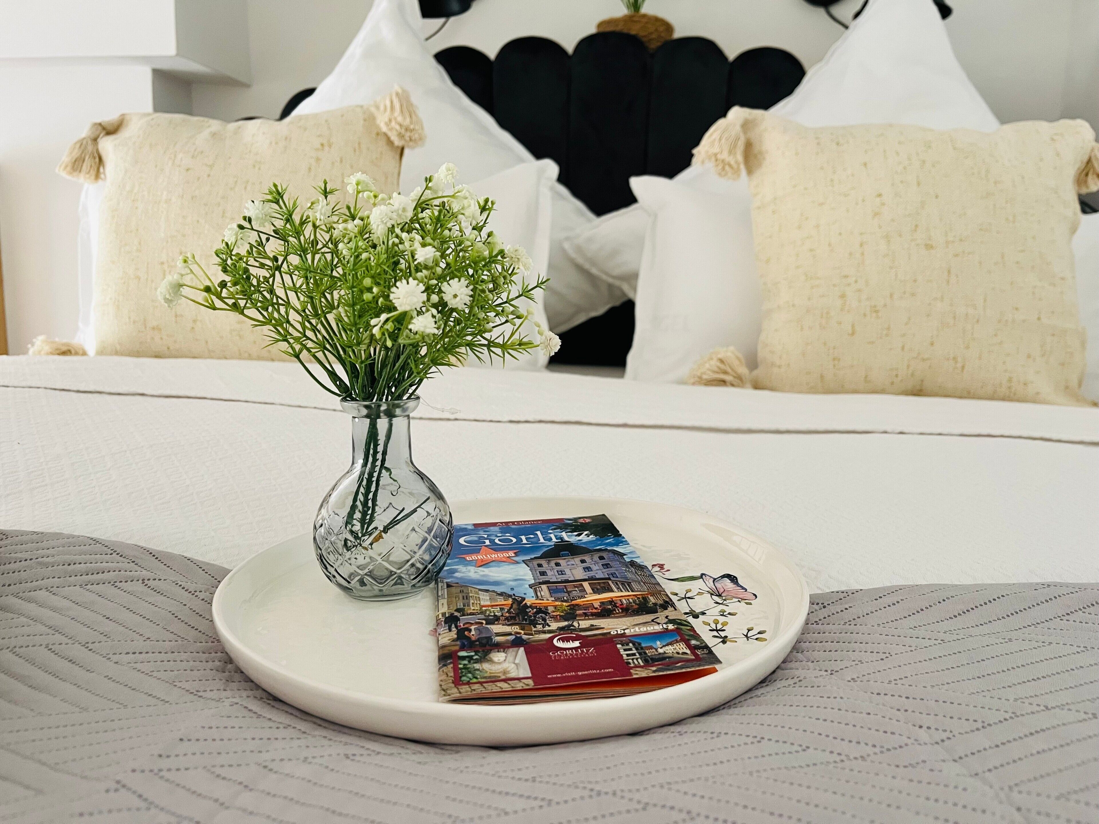 Ein Bett mit weißer Bettwäsche, eine transparente Vase mit weißen Blumen und ein Teller mit Reisebroschüren.