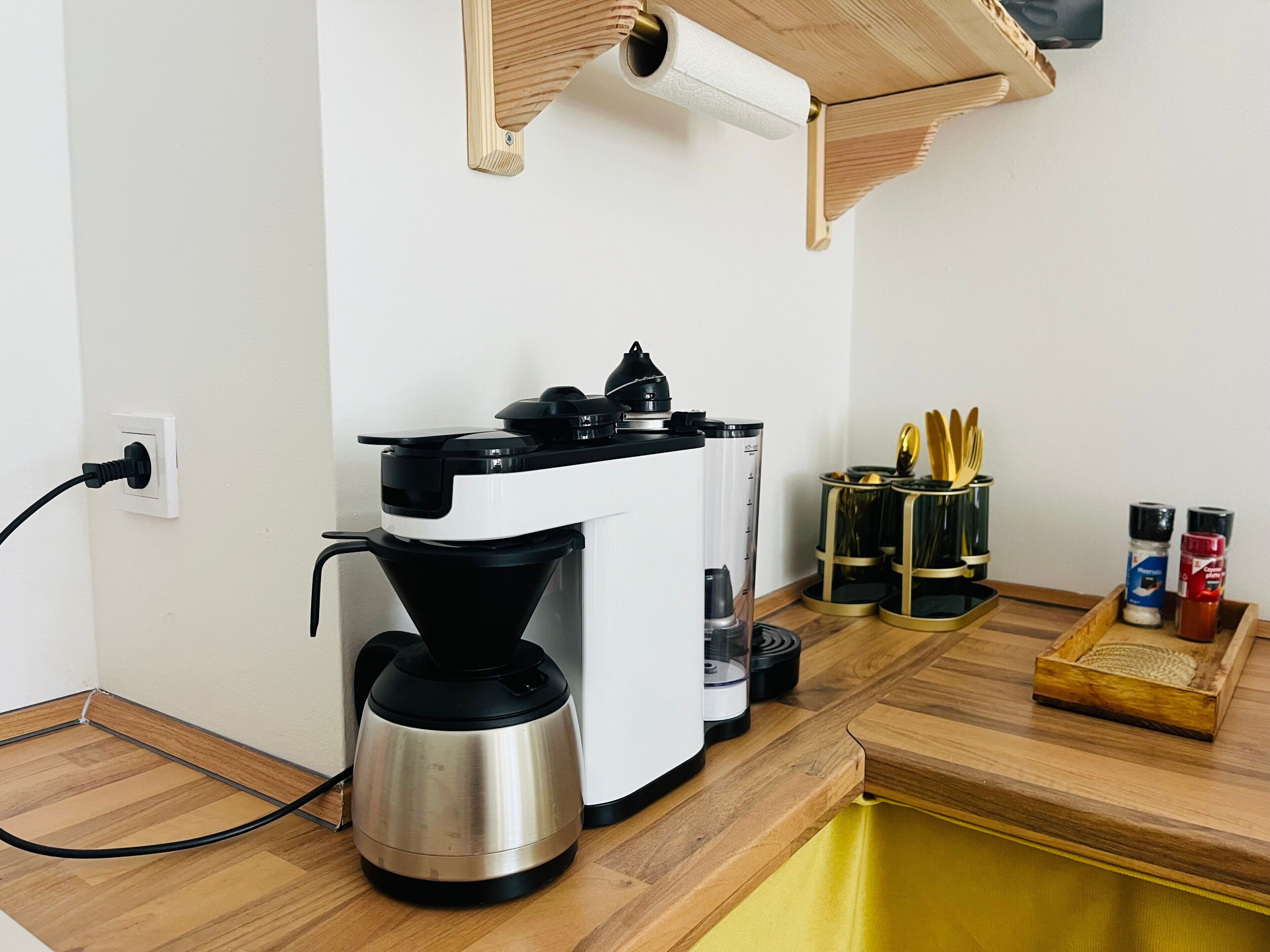 Une machine à café posée sur un plan de travail en bois, une prise électrique et un étagère à épices avec des récipients.