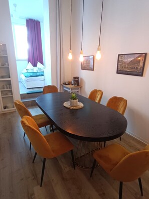 Un coin repas avec une table sombre, des chaises orange, des lampes suspendues, une machine à café et un tableau encadré au mur.