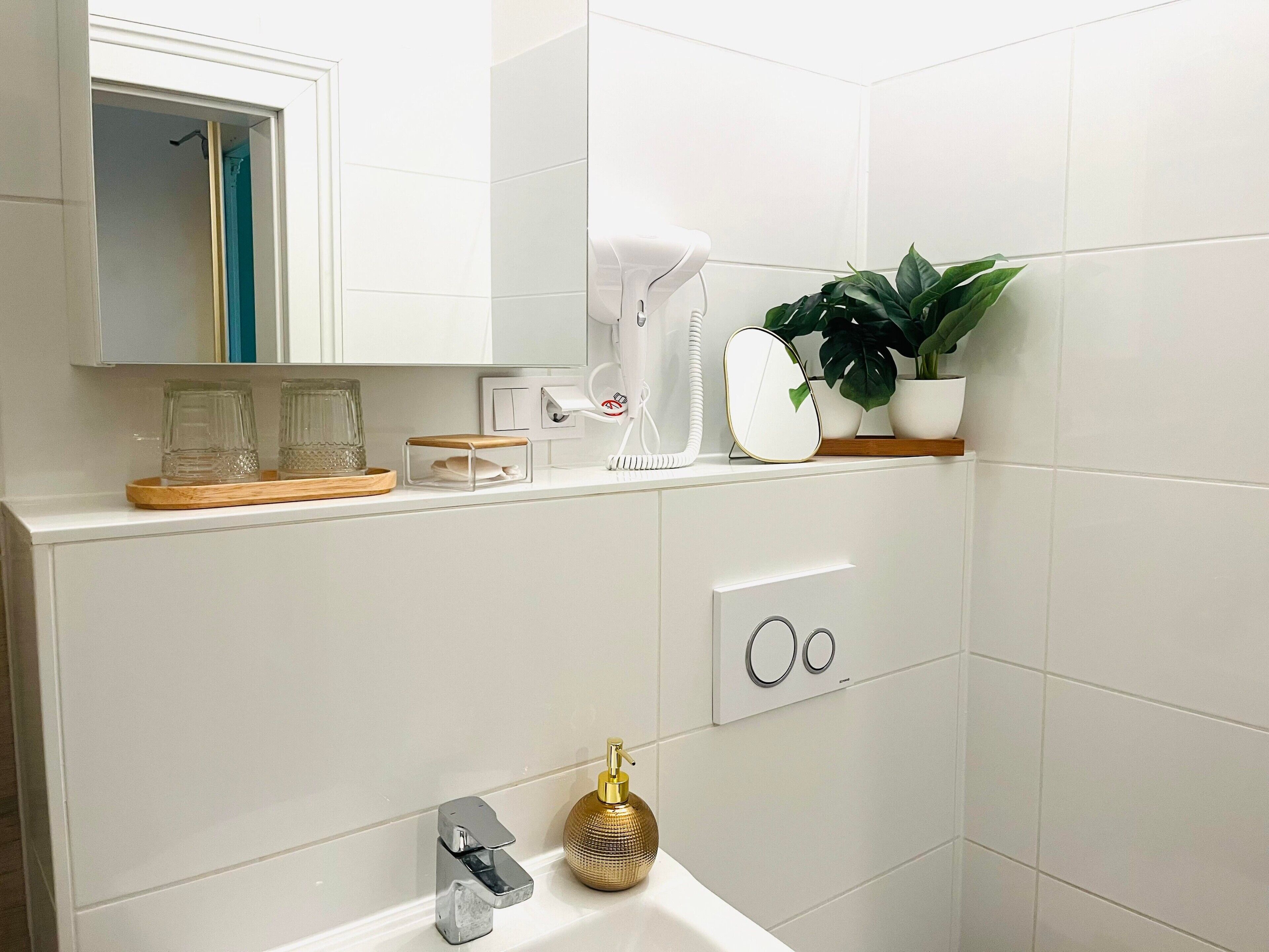 A modern bathroom with a white sink, a gold soap dispenser, a mirror, and a potted plant.