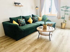 Un canapé vert agrémenté de coussins à motifs, une table basse ronde en bois sur laquelle est posée une plante, et une petite étagère sur laquelle se trouve un appareil photo ancien.