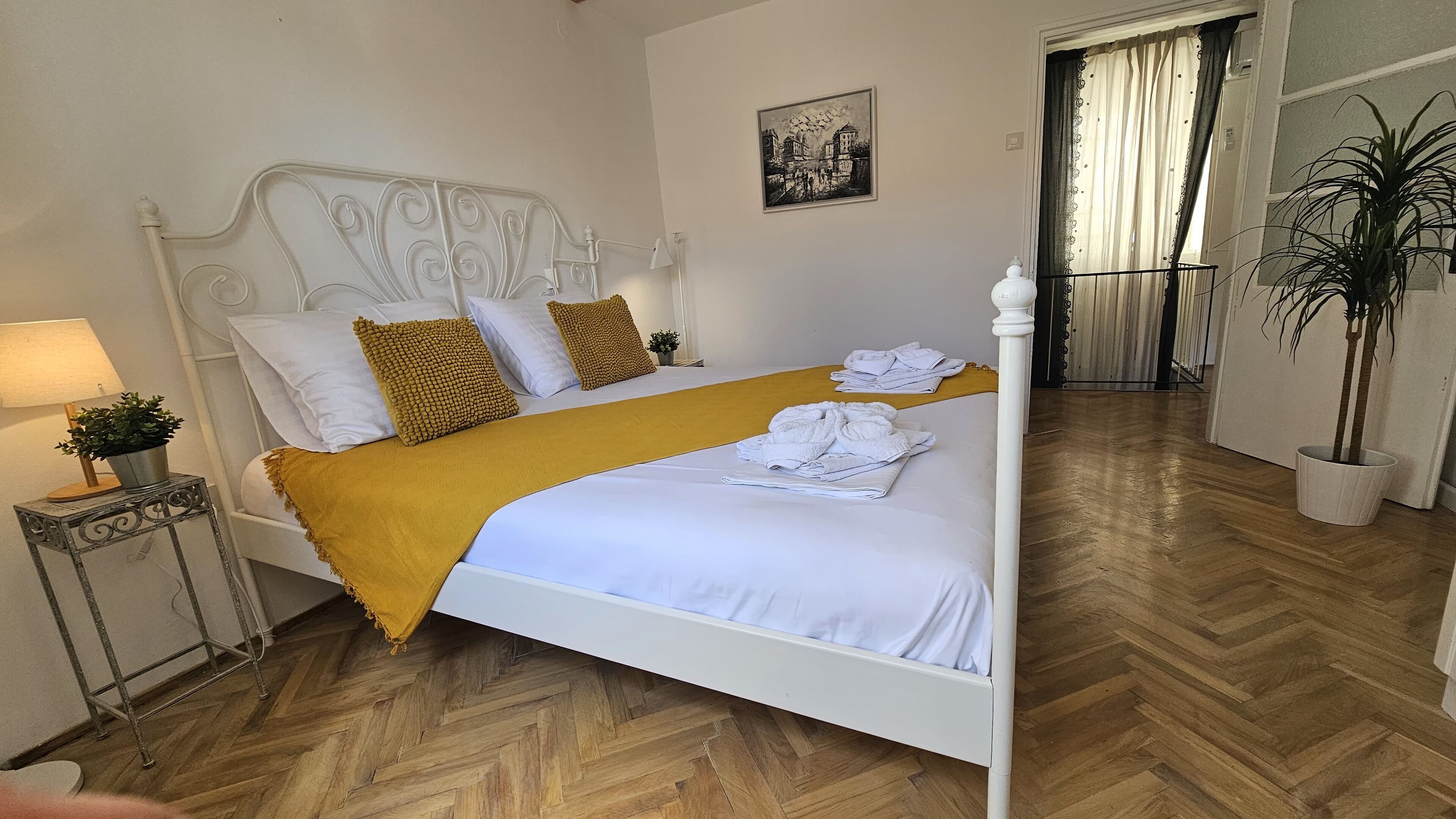 A neatly made bed with a white headboard, a yellow throw blanket, and a pair of towels on the bed. There is a small table with a lamp and a plant next to the bed.