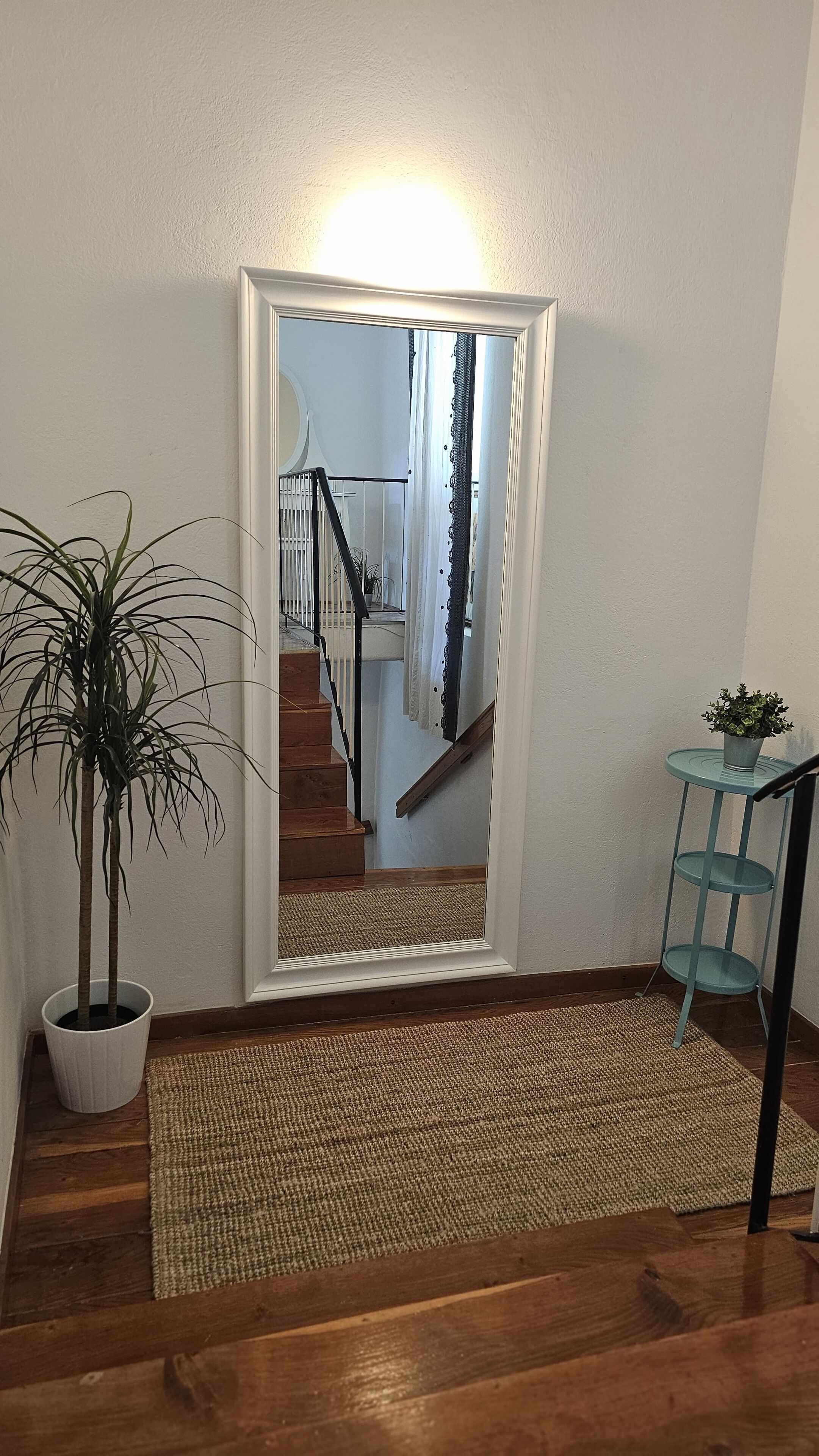 Un escalier avec une rampe en bois, un miroir à cadre blanc, une plante en pot et une étagère en métal bleue.