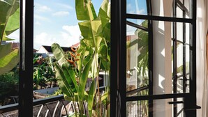 Une vue à travers une fenêtre dont le cadre est en métal, laissant entrevoir un feuillage verdoyant et quelques bâtiments à l’extérieur.