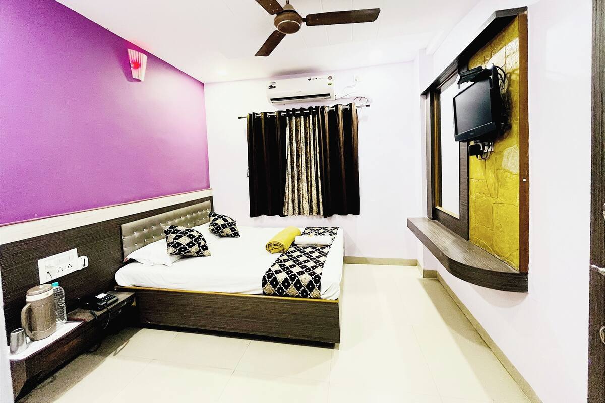 A hotel room with a bed, a ceiling fan, an air conditioner, a TV mounted on the wall, and a window with a view of the outside.