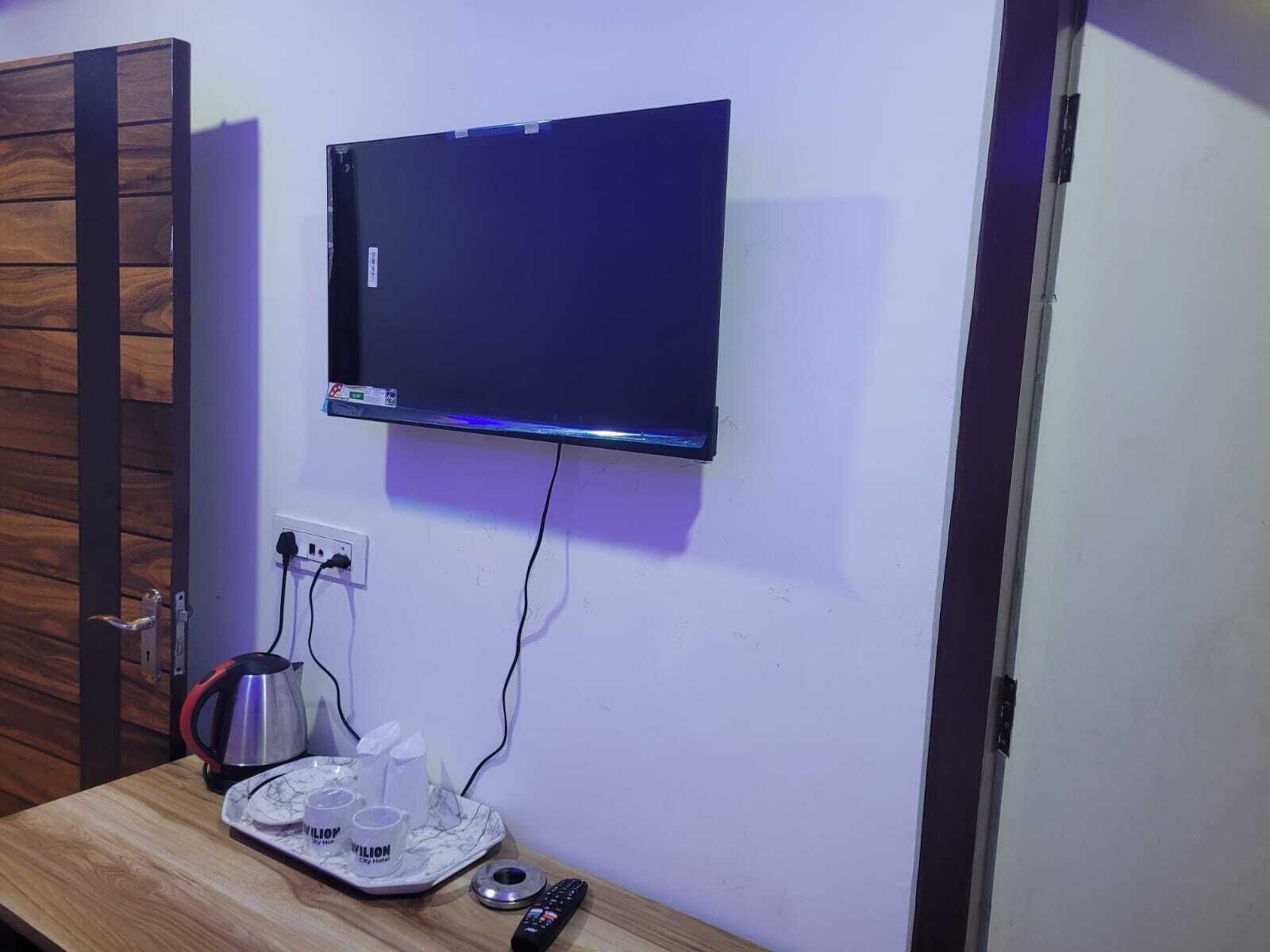 Un téléviseur à écran plat fixé au mur, une porte en bois, une petite table avec une bouilloire, des tasses et une télécommande.