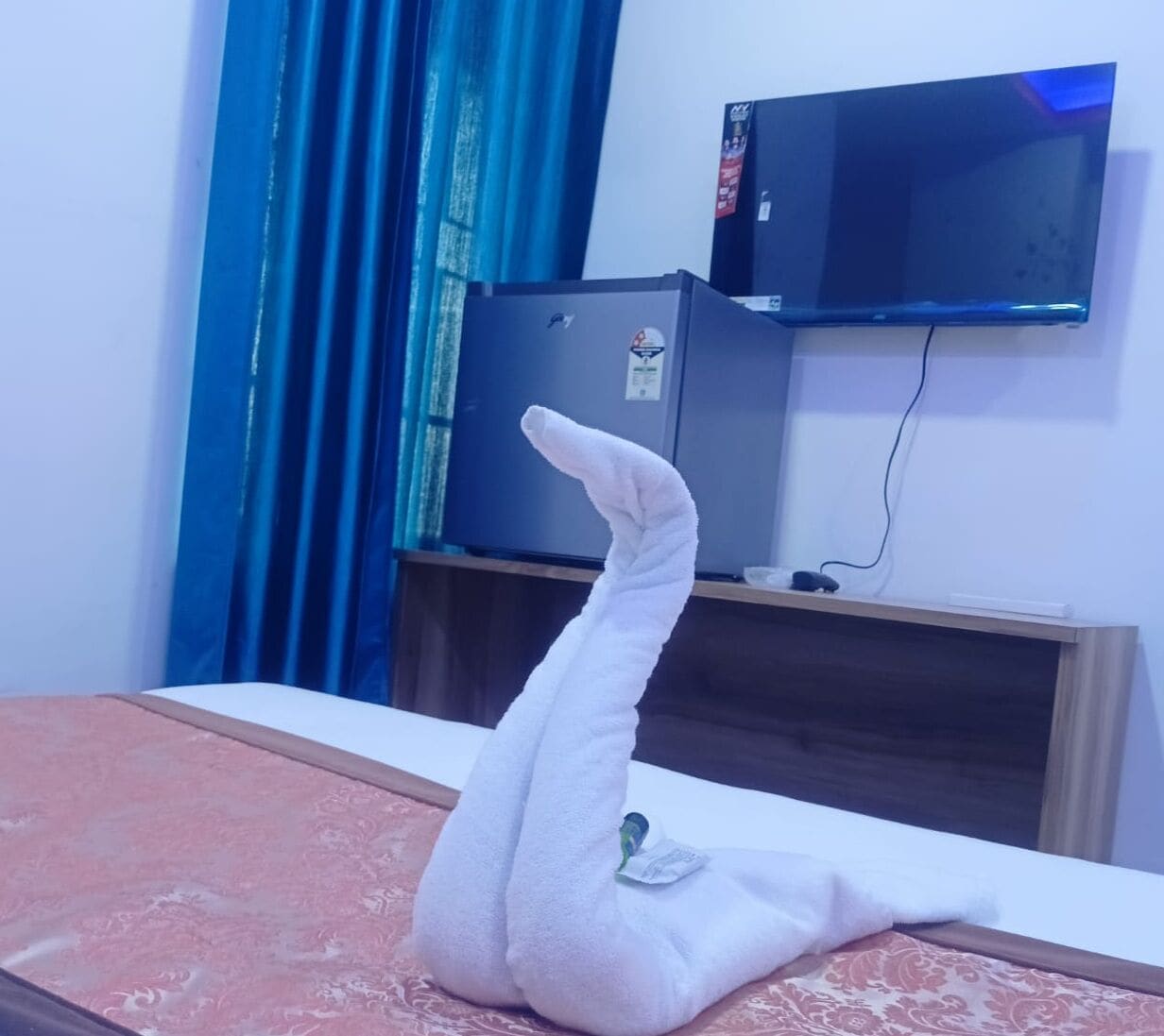 Een bed met een witte handdoek eroverheen, een aan de muur bevestigde televisie en een blauw gordijn.