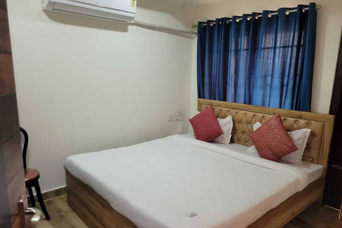 Quarto com cama de madeira, cabeceira, janela com cortinas azuis e ar condicionado na parede.