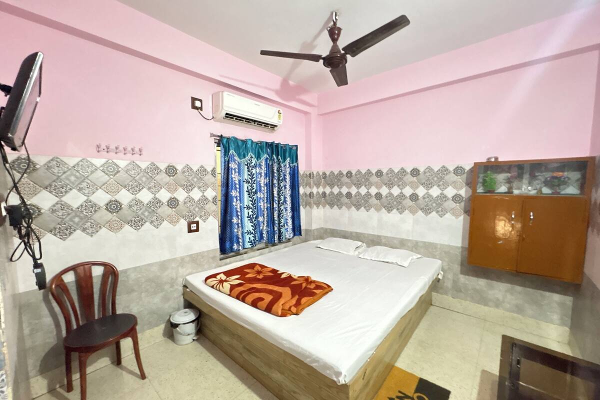 A bedroom with a bed, a ceiling fan, an air conditioner, a chair, a small trash bin, and a cabinet.
