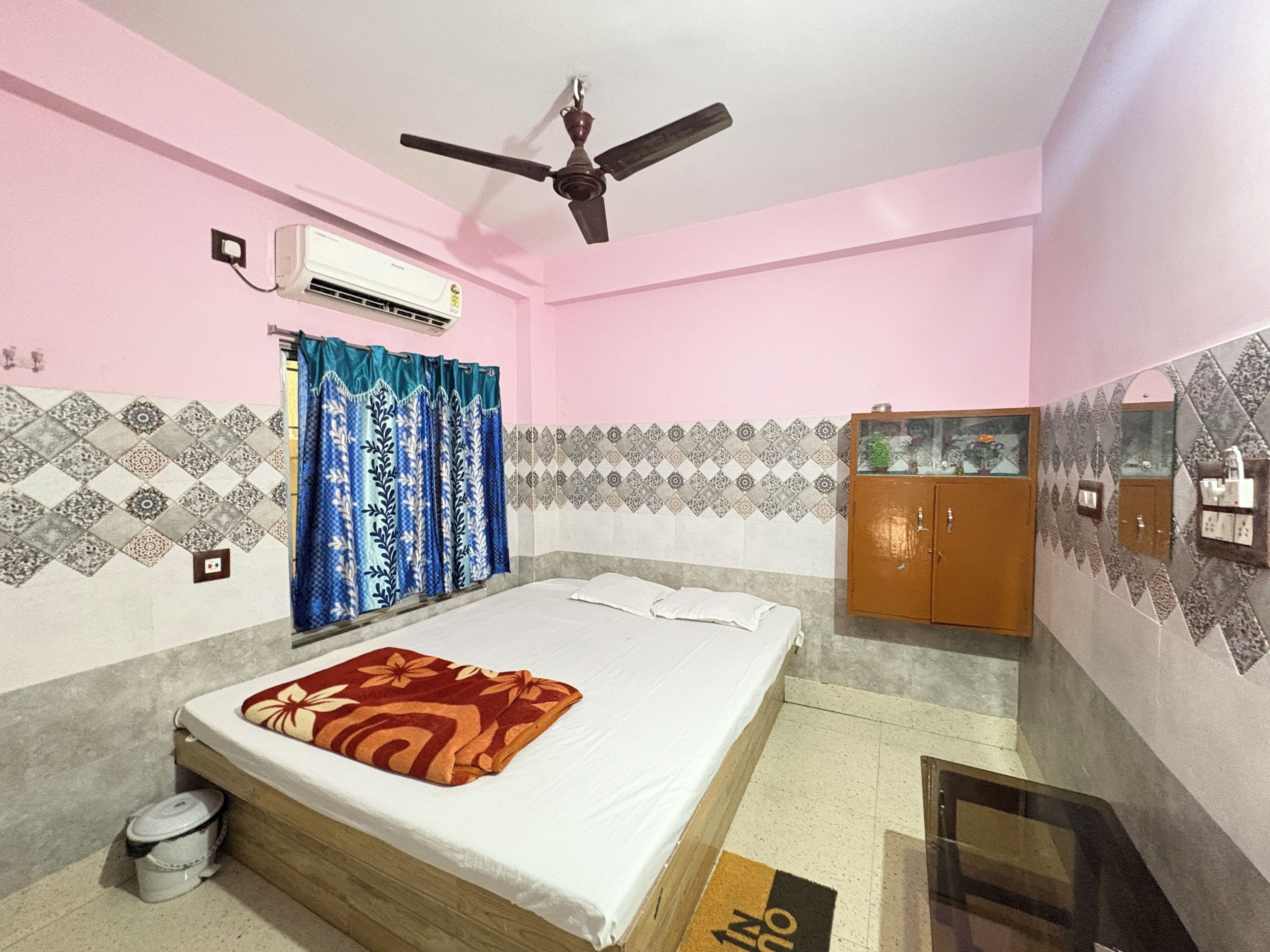 A bedroom with a bed, an air conditioner, a ceiling fan, patterned curtains, a cabinet, and a tiled wall.
