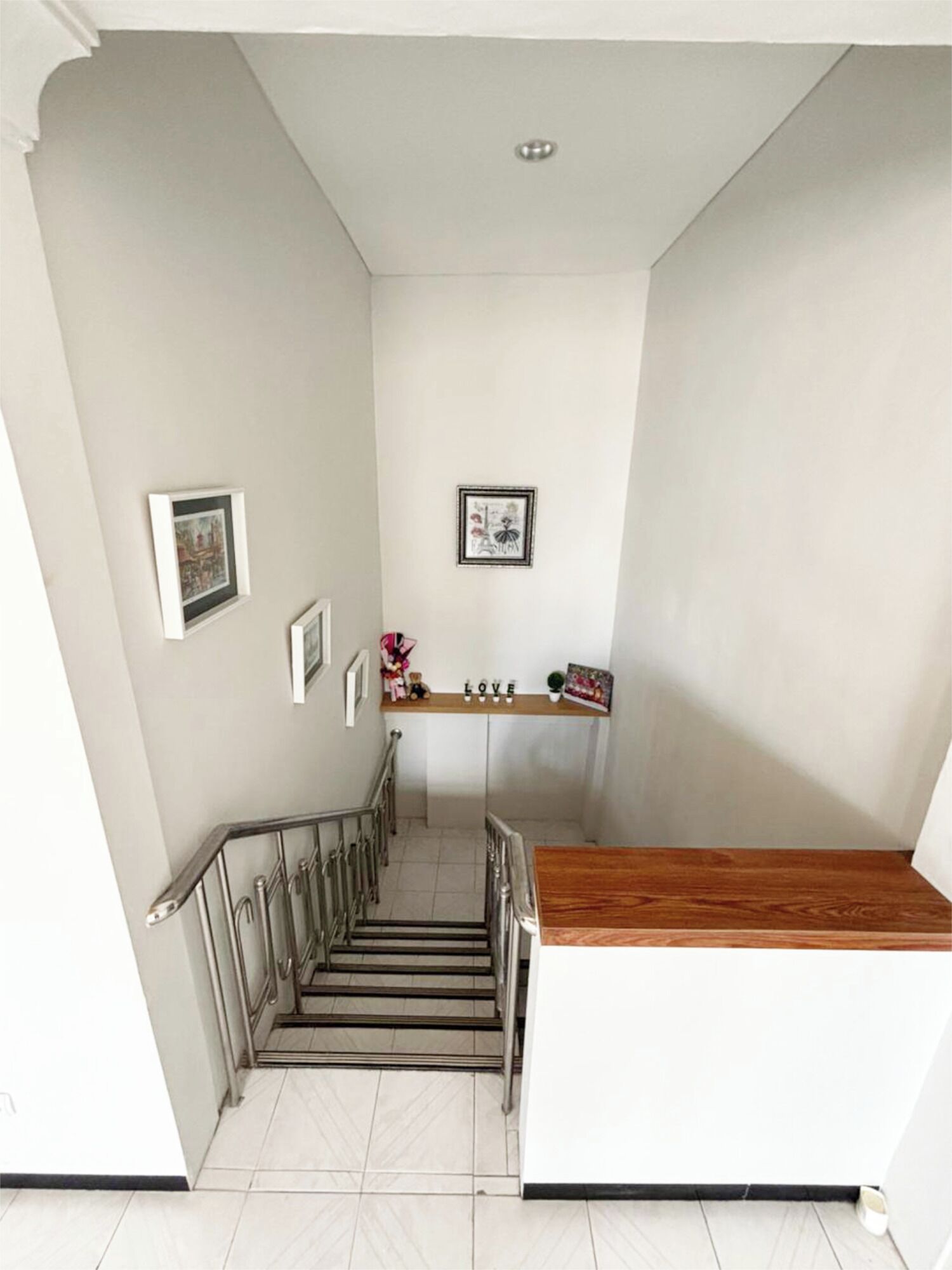 Una escalera con descansillo de madera y barandillas de metal, que conduce a una habitación con pared blanca y un pequeño estante con objetos decorativos.