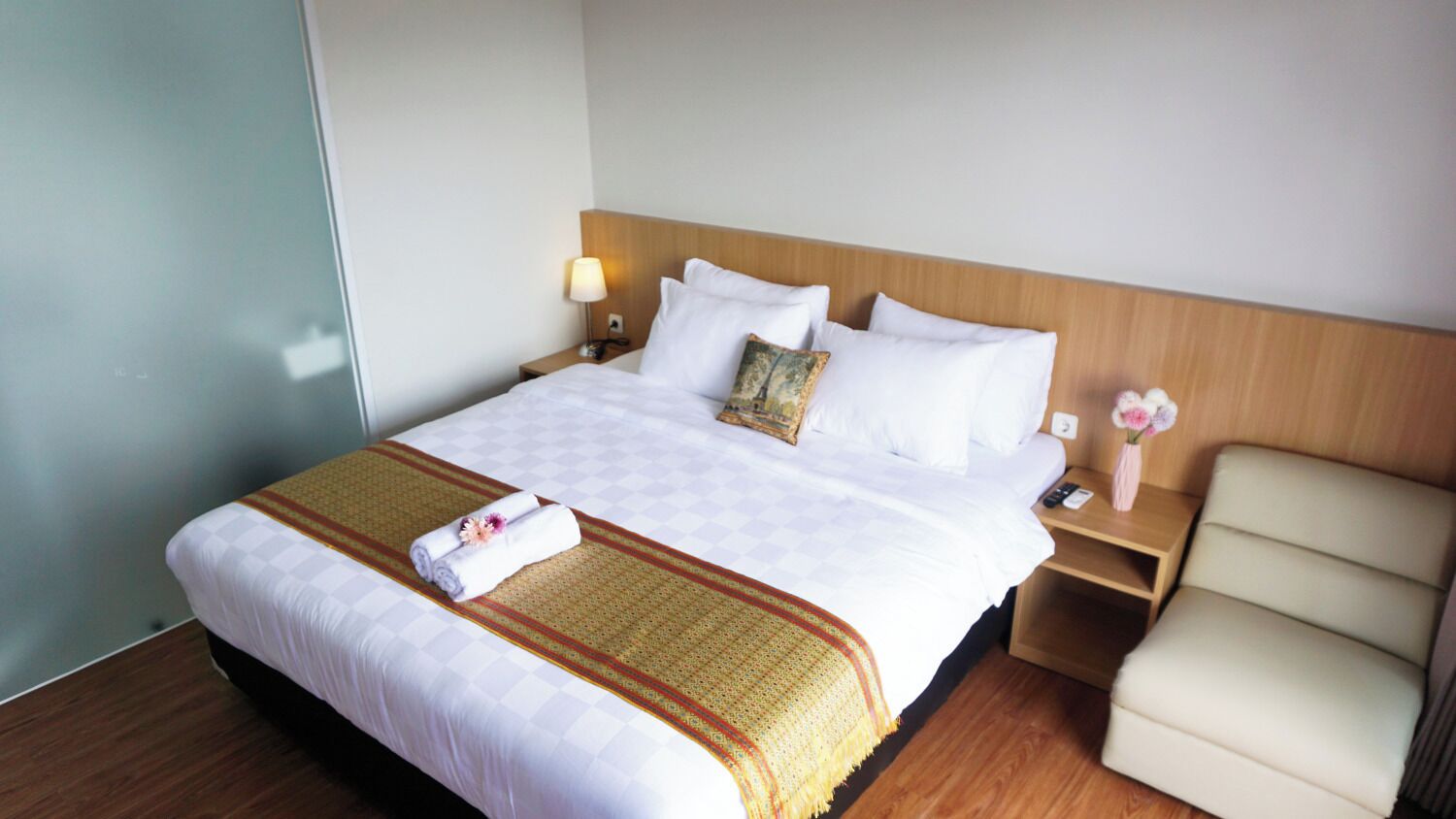 A hotel room with a large bed, a wooden headboard, a nightstand with a lamp, a chair, and a vase with flowers.