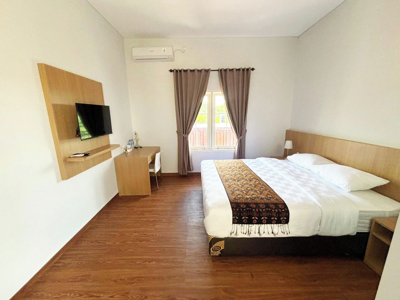 A hotel room with a bed, a desk, a chair, a TV mounted on the wall, and a window with curtains.