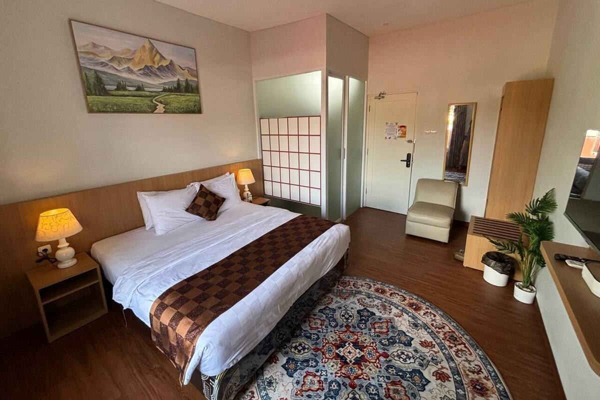 A hotel room with a bed, bedside lamps, a TV, a chair, a desk, a mirror, and a window.