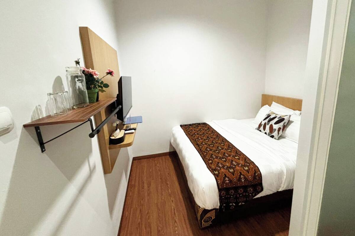 A small, neatly arranged hotel room with a bed, a TV, a shelf with a vase of flowers, and a wooden floor.