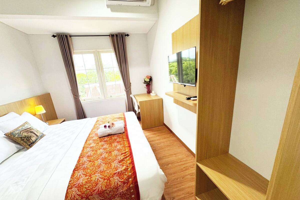 A hotel room with a bed, a TV mounted on the wall, a small desk, and a window with curtains.