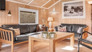 A cozy living room with wooden walls and ceiling, a large window, two leather sofas, a wooden coffee table, and a framed photograph on the wall.