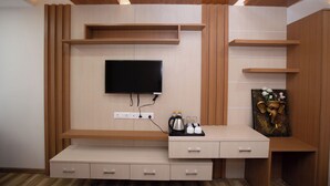 A modern living room with a flat-screen TV mounted on a wooden wall, a coffee maker, and a small shelf with decorative items. - Hotel Ambrosia (Bhimeshwar)