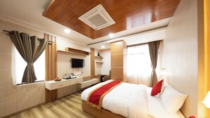 A modern hotel room with a wooden ceiling, a large bed with white bedding and red accents, a flat-screen TV, a desk with a chair, and a window with curtains. - Hotel Ambrosia (Bhimeshwar)