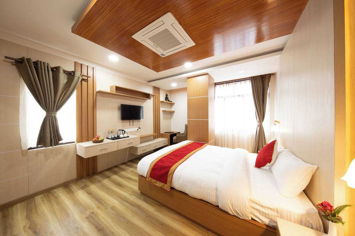 A modern hotel room with a wooden ceiling, a large bed with white bedding and red accents, a flat-screen TV, a desk with a chair, and a window with curtains.