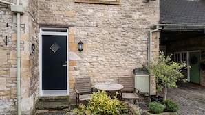 Un bâtiment en pierre avec une porte noire, une petite table ronde et deux chaises à l’extérieur.