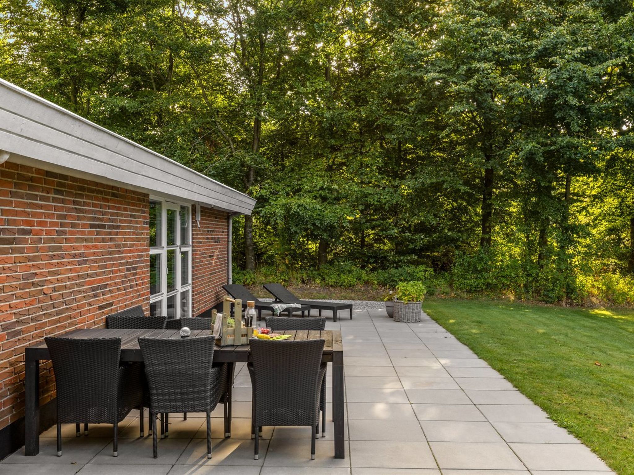Une terrasse avec un mur en briques, des meubles d’extérieur et une zone herbeuse agrémentée d’arbres.