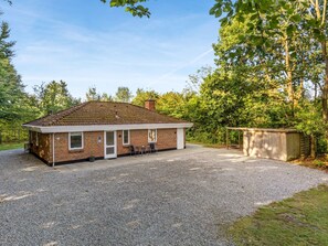 A single-story brick building with a gravel driveway, a small patio with outdoor seating, and a wooden shed in a wooded area.