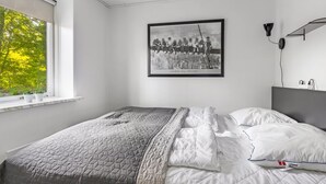 Ein Schlafzimmer mit Bett, Fenster mit Blick auf Bäume, gerahmtes Bild an der Wand und Nachttisch mit Lampe.