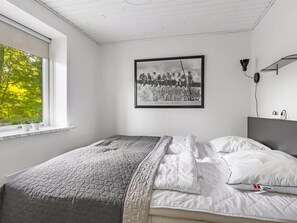 Una camera da letto con un letto, una finestra vista alberi, un quadro appeso al muro e un comodino con una lampada.