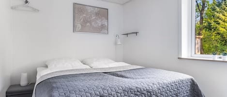 A bedroom with a bed, a bedside table, a wall-mounted light, and a window with a view of trees.