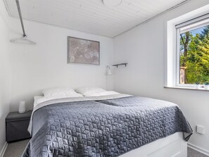 A bedroom with a bed, a bedside table, a wall-mounted light, and a window with a view of trees.
