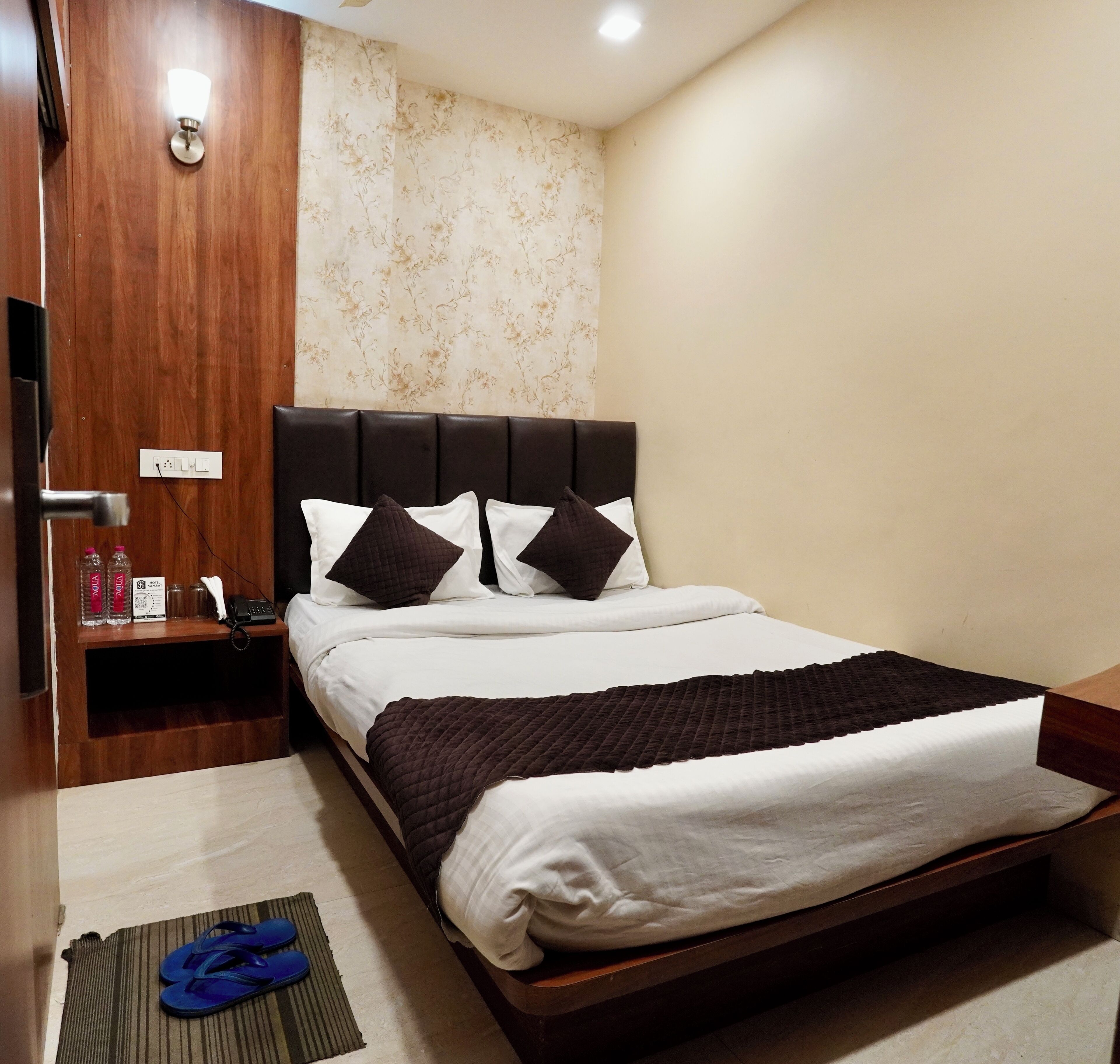 A neatly made bed with a dark headboard, a bedside table with a lamp, and a wall with floral wallpaper.