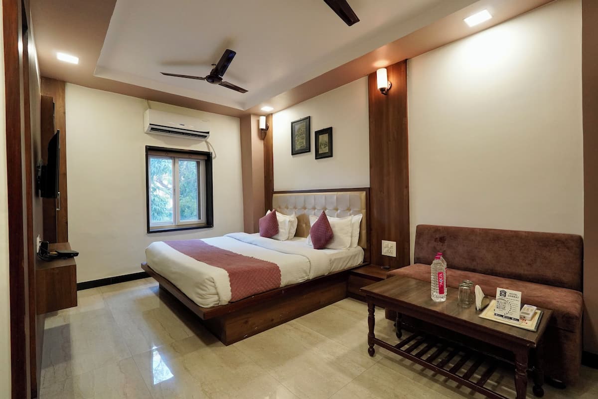 A hotel room with a bed, a sofa, a table, a ceiling fan, an air conditioning unit, and a window.