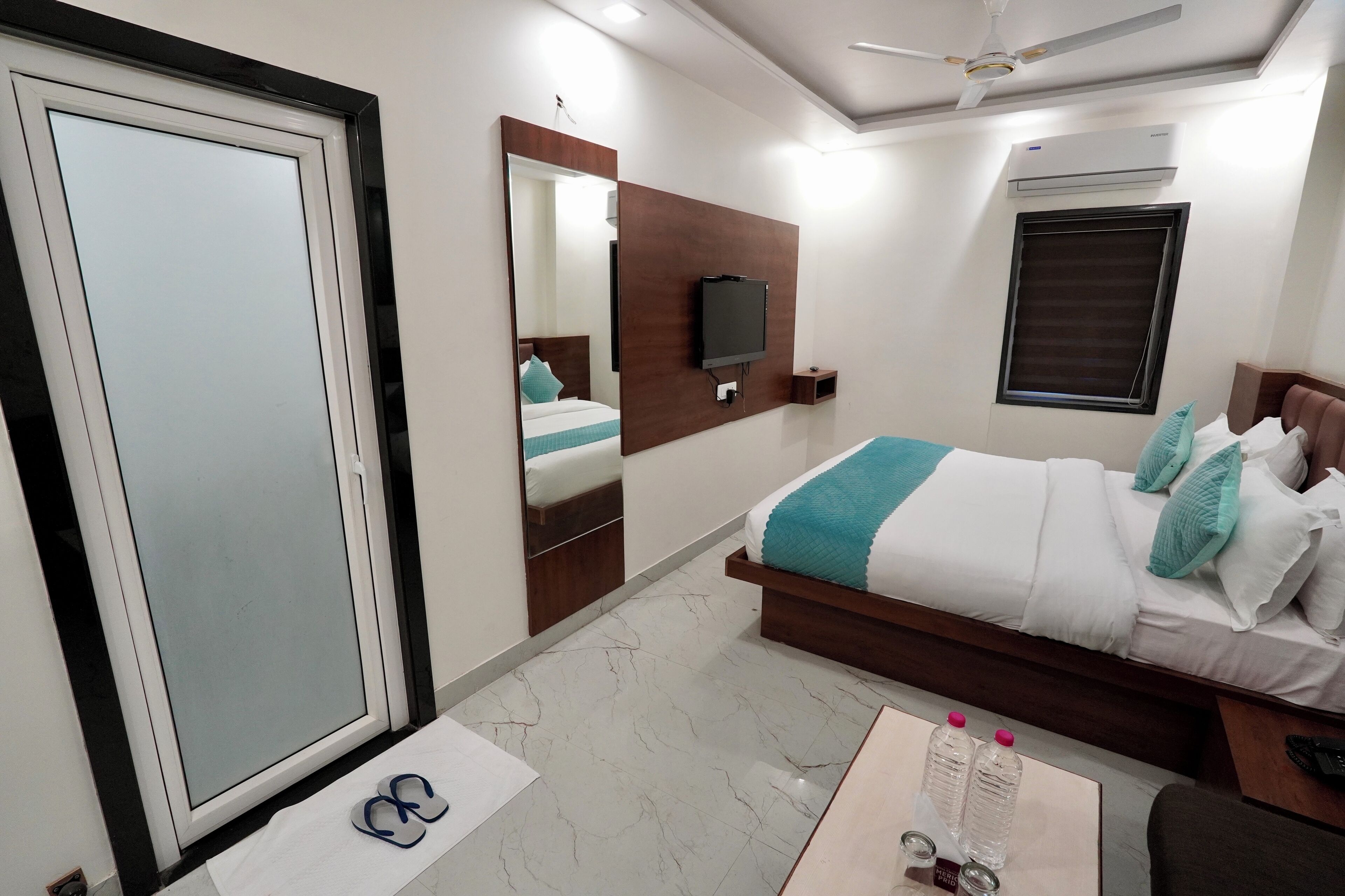 A modern hotel room with a large wardrobe, a bed with blue and white bedding, a flat-screen TV mounted on the wall, and an air conditioning unit.