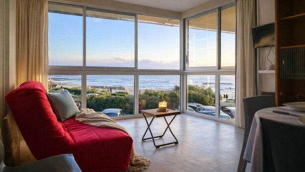 A room with large windows offering a view of the beach and ocean. There is a red sofa with a blanket, a small table with a candle, and a TV on a wooden shelf. - Sea-facing apartment - 5 people (Les Sables-d´Olonne)