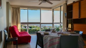 A dining area with a table set for four, a red chair, and a view of greenery through large windows. - Sea-facing apartment - 5 people (Les Sables-d´Olonne)