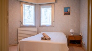 A bedroom with a bed, two windows with curtains, a bedside table with a lamp, and a wall-mounted picture. - Sea-facing apartment - 5 people (Les Sables-d´Olonne)