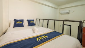 A hotel room with a neatly made bed, two pillows with the hotel's logo, a rolled towel, and a wall-mounted air conditioner. - CQA Living By ARBA (Jakarta)