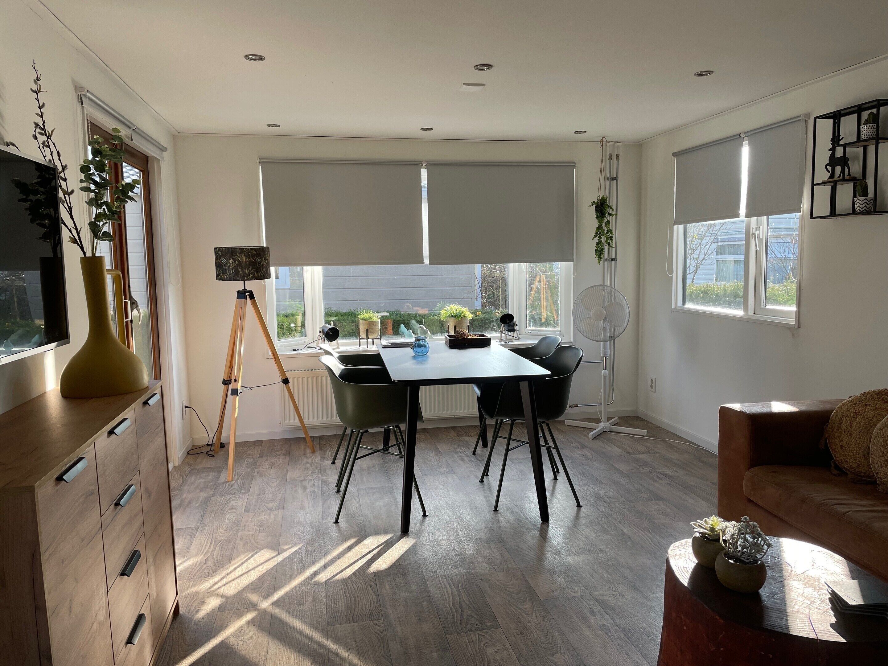A modern living room with a dining area, featuring a large window, a standing fan, and a sofa.