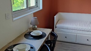 A compact room with a bed, a dining table with chairs, a lamp, and a window with a view of greenery.