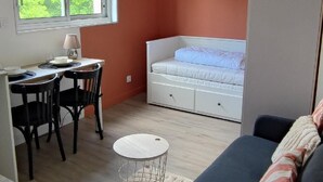 A compact room with a bed, a desk with chairs, a small table, and a window with a view of greenery.