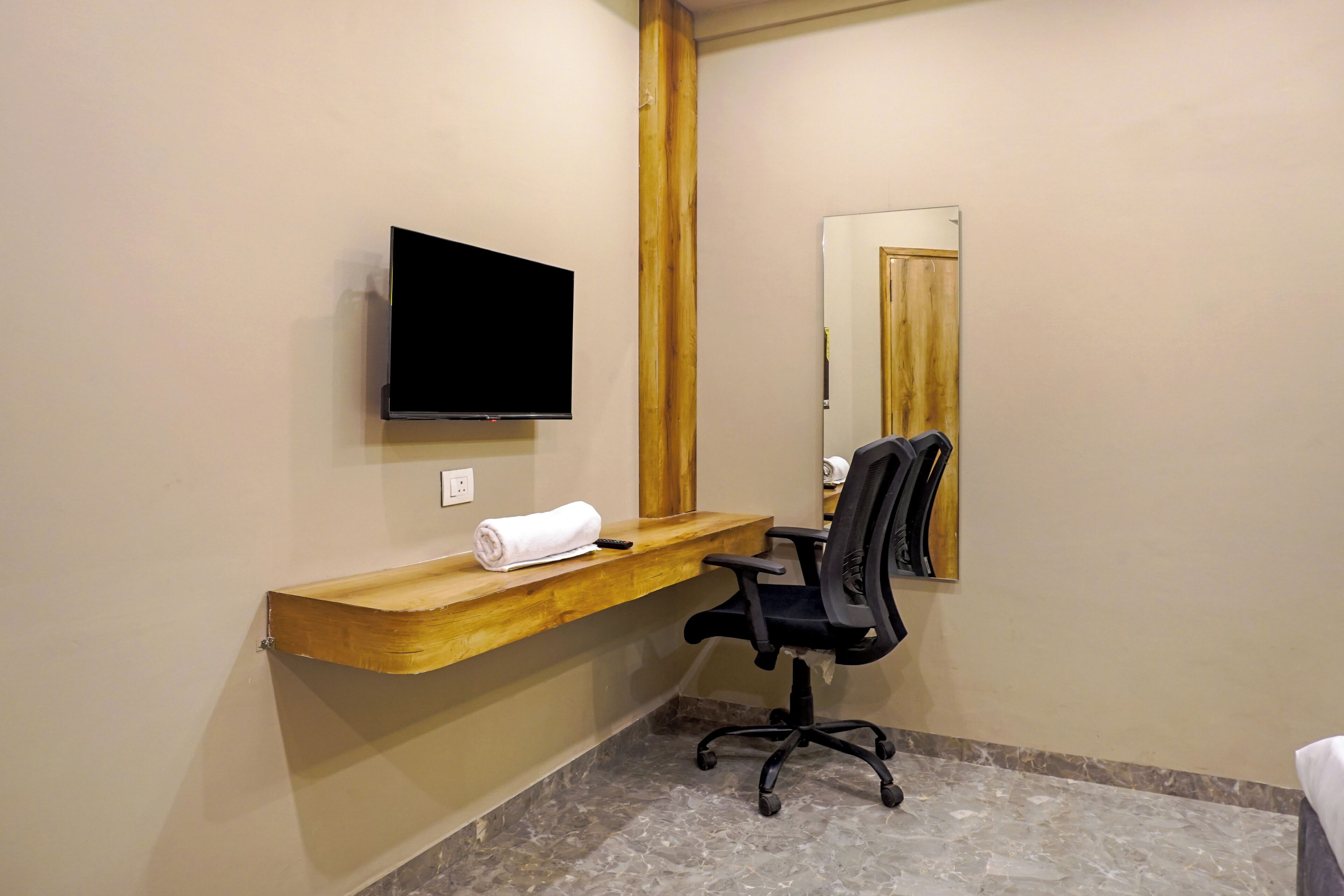 Un baño con tocador de madera, una silla de oficina negra, un televisor de pantalla plana montado en la pared y un espejo sobre el tocador.