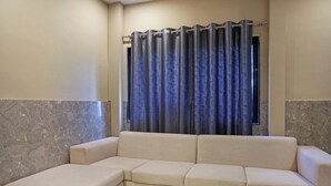 A beige sectional sofa in a corner with a window and curtains. - Hotel Nest Inn (Mumbai)