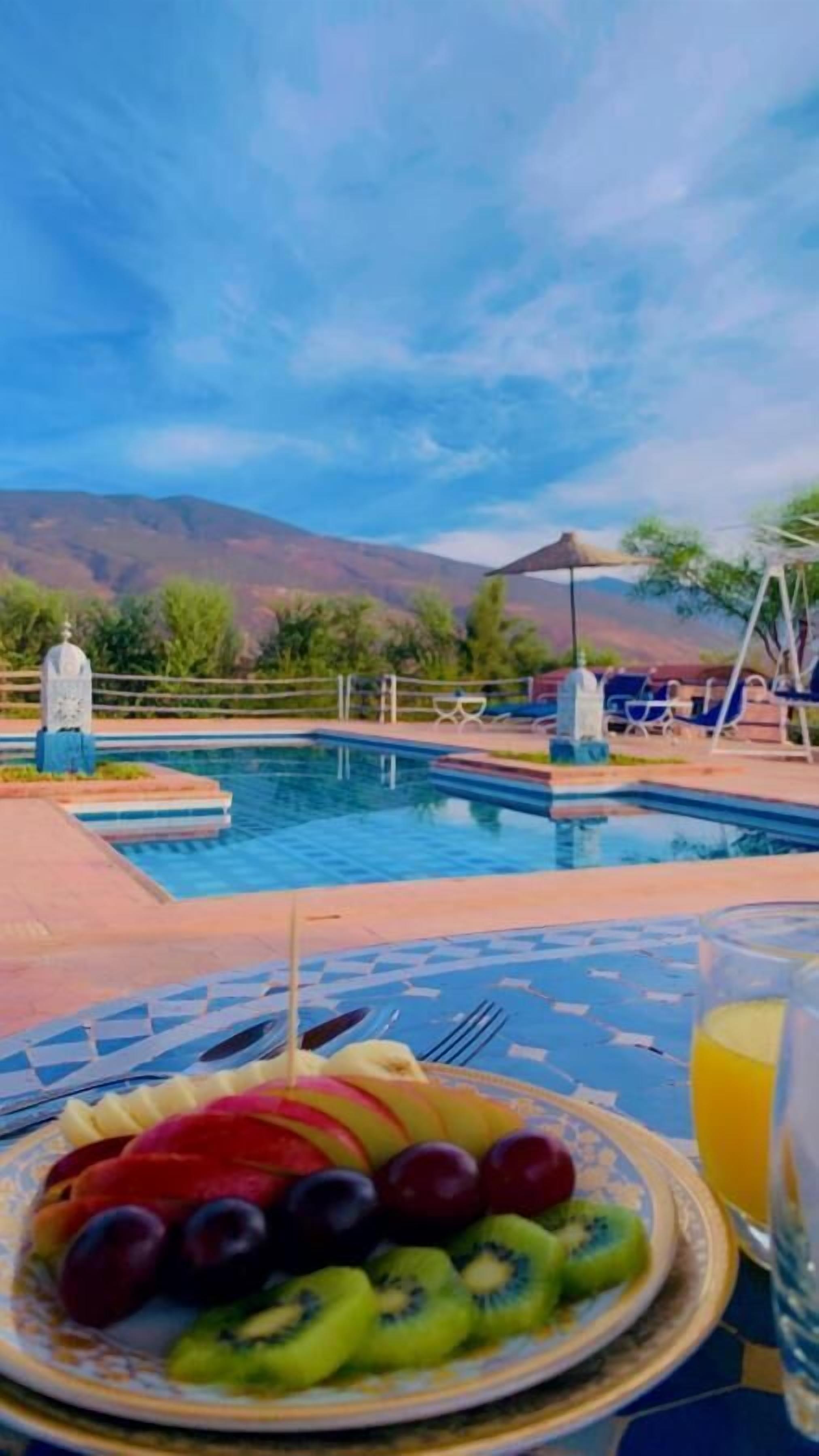 Ein Tisch am Pool mit einer Obstplatte, einem Glas Saft und Blick auf Pool und Berge.