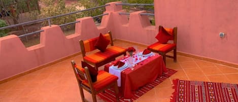 Una terraza con una mesa preparada para dos, dos sillas de madera con cojines rojos, un mantel rojo y una alfombra estampada.