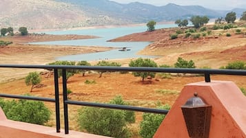Une vue depuis un balcon donnant sur une rivière où naviguent des bateaux, le tout entouré d’un paysage aride et rougeâtre, parsemé de quelques rares zones de végétation.