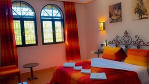 Ein Zimmer mit einem Bett, zwei Fenstern, einem Tisch und einer Wandlampe.