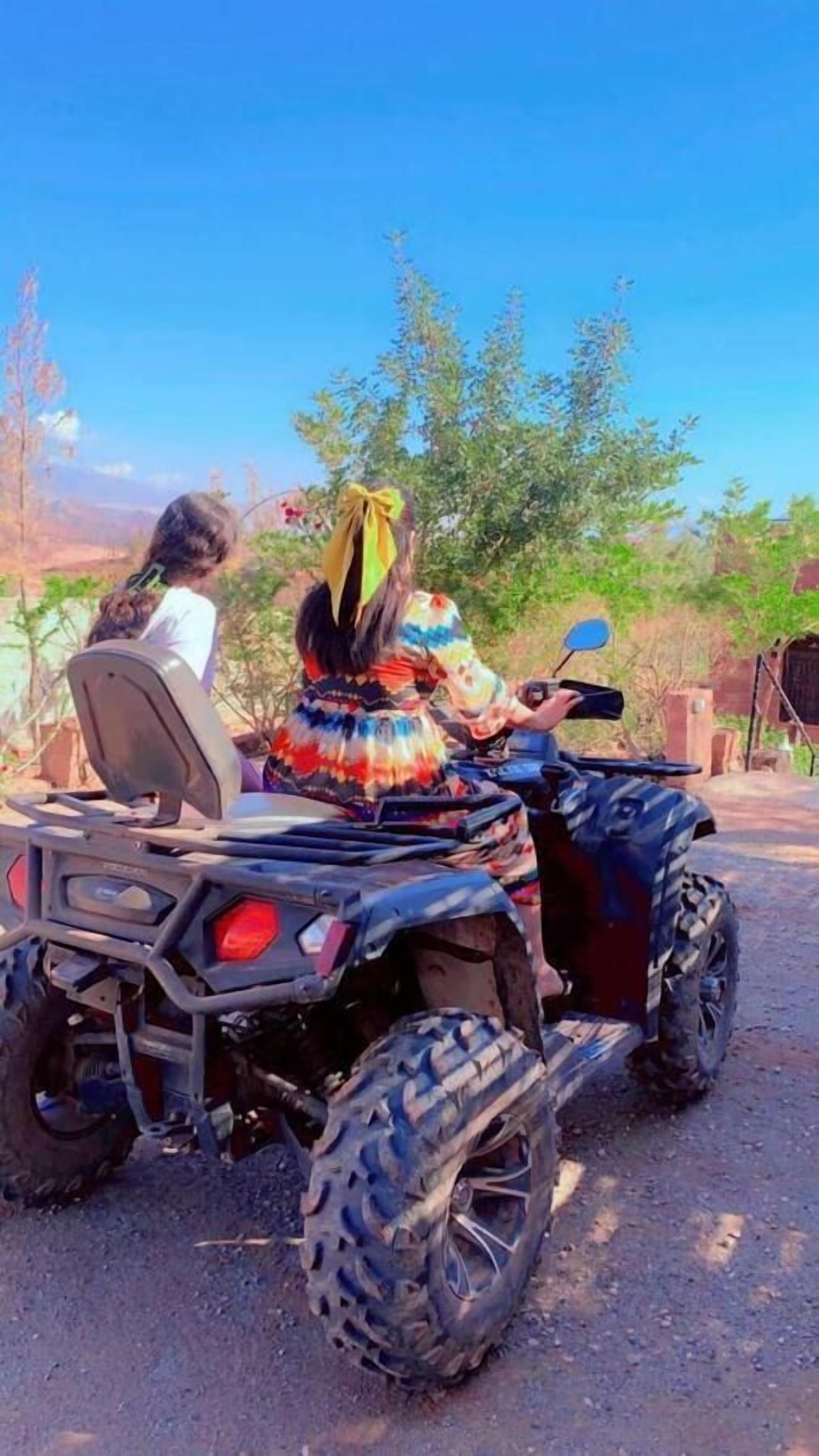 Deux personnes sur un quad, dans un paysage rural, avec des arbres et un ciel dégagé.